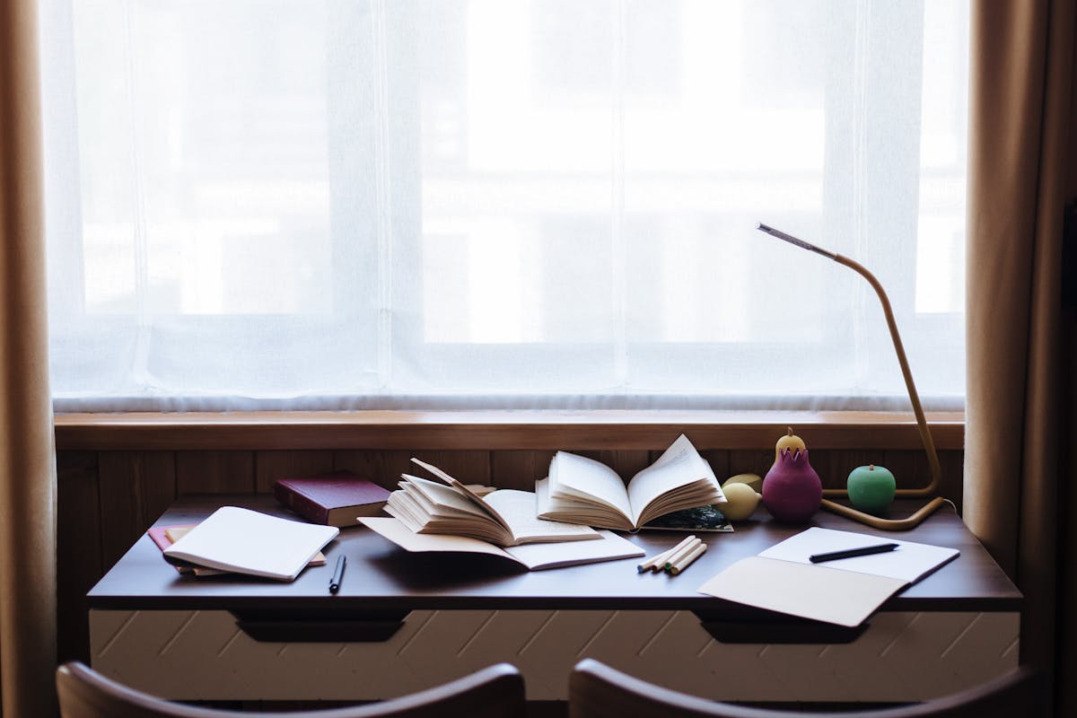 A well-lit study area with open books, notebooks, and a lamp on a desk by the window.